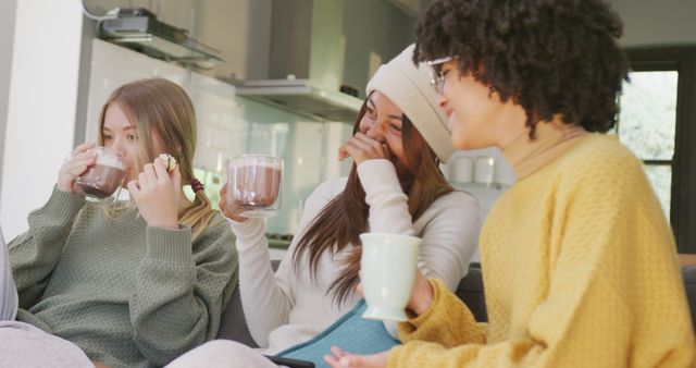 Happy diverse teenager girls sitting on sofa, drinking cocoa and laughing. Spending quality time, lifestyle, friendship and adolescence concept.