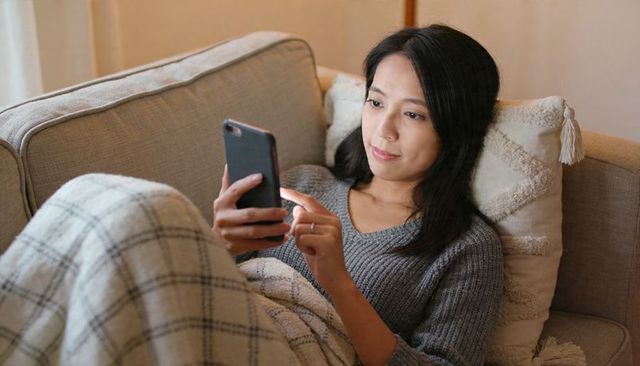 Asian woman relaxing on sofa scrolling smartphone under plaid blanket with tassel pillow