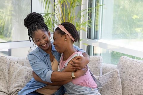 African mother and teen daughter hugging and smiling on sunlit modern living room sofa