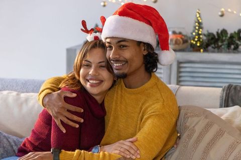 Diverse couple cuddling on sofa wearing santa hat and knit sweaters during cozy christmas evening