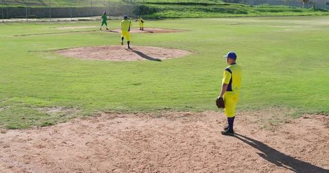 Diverse male baseball players standing on sunlit field holding gloves wearing yellow uniforms