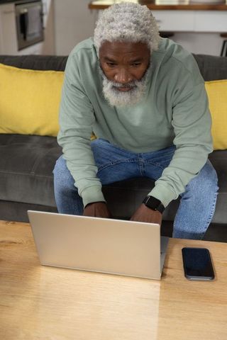 Senior african american man working on laptop at home on sofa, focused remote work