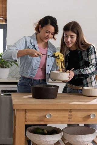 Two friends serving pasta into ceramic bowls at sunlit wooden kitchen island