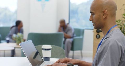 Indian healthcare worker working on laptop in clinic lounge with colleagues in scrubs