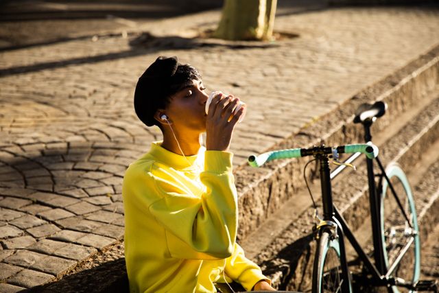 Side view of a fashionable biracial transgender in the street, leaning on a wall, drinking coffee, listening to music on earphones, with a bike standing by him