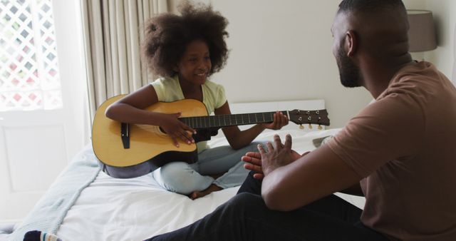 This image shows a father and daughter enjoying playing the acoustic guitar together in a cozy home environment. The daughter is sitting on the bed holding the guitar, while the father is attentively looking at her with a smile. This image is perfect for depicting family bonding time, music lessons at home, educational content about music, or promoting family-oriented services and products.