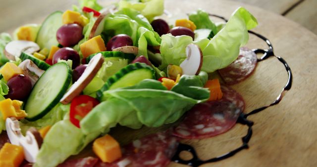 Fresh vegetable salad featuring grapes, cheese cubes, cucumber slices, lettuce, and mushroom slices arranged on a wooden plate with a salami base and balsamic glaze. Ideal for promoting healthy eating, vegan menus, gourmet meals, and food blogs.