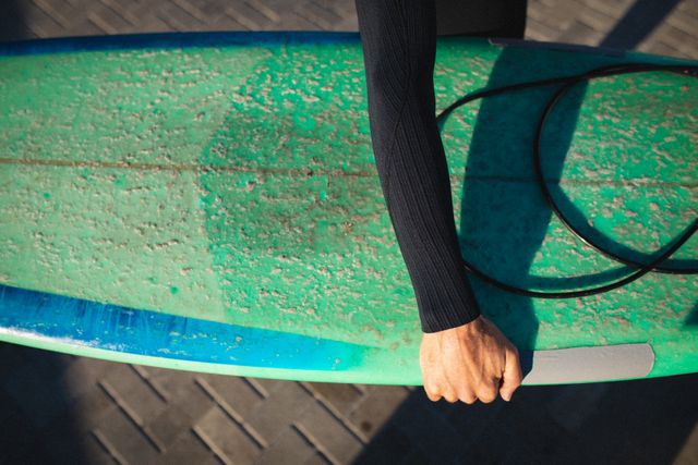 Cropped view of a male surfer holding a turquoise surfboard on a sunny day. Ideal for promoting surfing activities, beach vacations, water sports, and active lifestyles. Perfect for use in travel brochures, sports magazines, and adventure blogs.