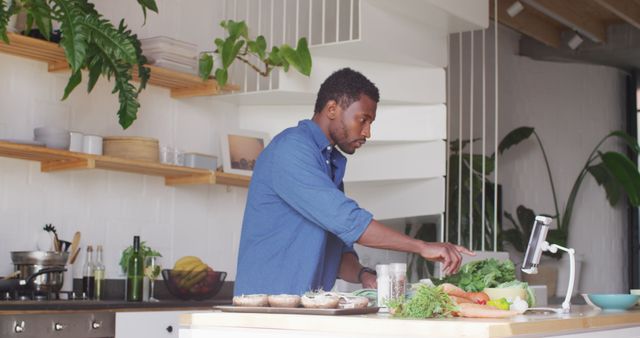 A man is preparing a healthy meal in a modern kitchen with a focus on fresh vegetables and green plants around. This image can be used for lifestyle blogs, healthy cooking promotions, kitchen design inspirations, and advertisements for organic food products.