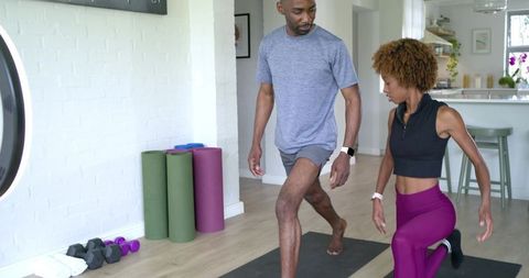 Diverse couple lunging together in bright modern kitchen workout with mats and dumbbells