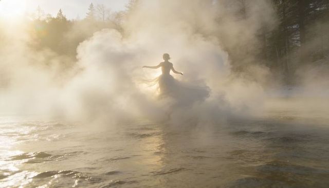 Backlit woman in flowing gown wading through misty lake at golden hour