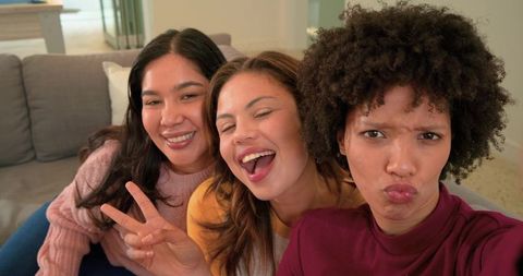 Diverse female friends taking joyful selfie on couch laughing and making peace sign