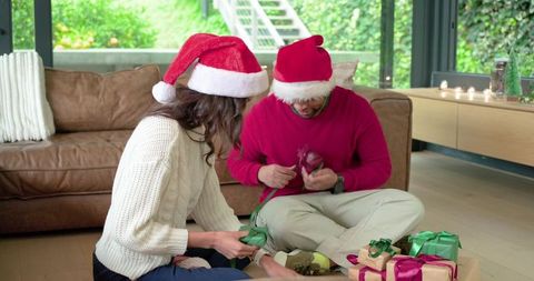 Couple wrapping christmas gifts on living room floor wearing santa hats and cozy sweaters