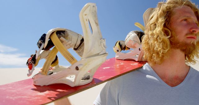 Young man carrying snowboard over his shoulder, standing in sunny desert under bright blue sky. Ideal for themes related to extreme sports, outdoor activities, adventure, and active lifestyle content.