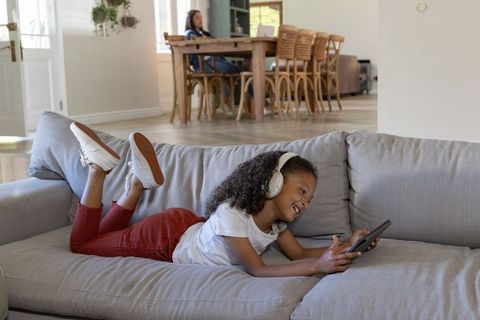 Smiling African American girl lying on couch using tablet with headphones in open-plan home