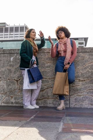 Female friends chatting and shopping at urban plaza holding bags and scarves