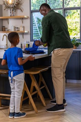 African american father packing son's blue backpack at kitchen island morning routine