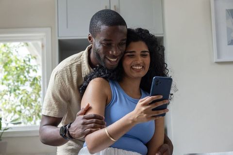 Smiling diverse couple embracing and using smartphone in bright kitchen, natural light