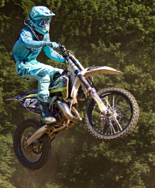 Rider wearing protective gear performing high jump on motocross circuit, showcasing skill and control. Useful for themes related to motocross events, extreme sports, outdoor activities, and adventure sports marketing materials.