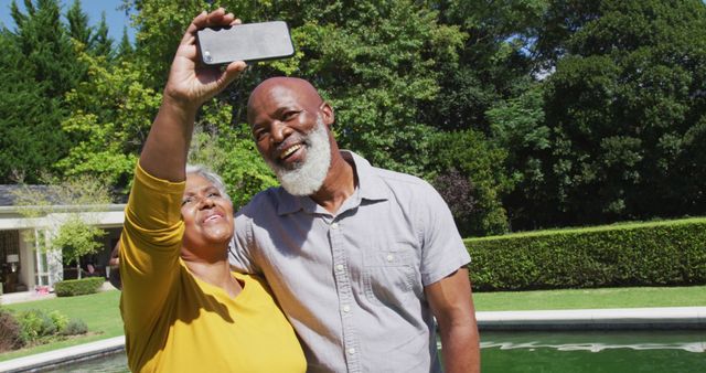 Happy senior african american couple using smartphone taking selfie by swimming pool in sunny garden. retirement, technology, senior lifestyle and leisure time.