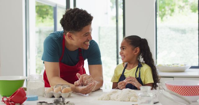 Father and daughter are cooking together in a modern kitchen. They are smiling and enjoying the process of baking with ingredients like flour, dough, and eggs. This image can be used in advertisements for family-oriented products, cooking classes, or kitchen appliances. It captures a warm, joyful moment between parent and child.