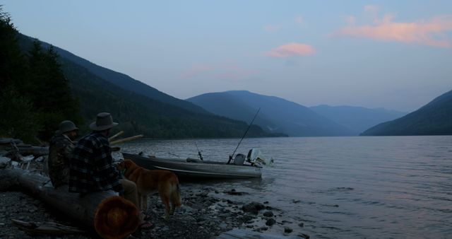 Two middle-aged Caucasian men and a dog are relaxing by a lake at dusk, with a boat and fishing gear nearby, with copy space. A serene outdoor setting is captured as the men enjoy a peaceful fishing trip in the wilderness.