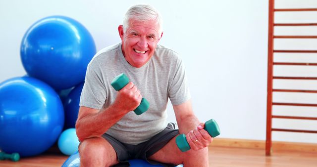 Elderly man lifting hand weights sitting on exercise ball in the gym