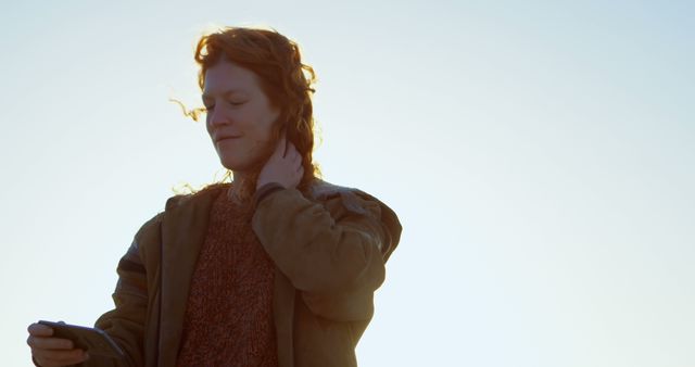 Person standing outdoors in early morning light, red hair gently blowing in the wind. Looking at smartphone, wearing jacket and casual attire. Great for themes of technology use in natural settings, enjoyment of morning light, or casual outdoor activity.