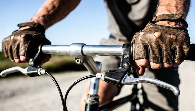 Gripping leather-gloved hands steering vintage bicycle handlebars on sunlit gravel road