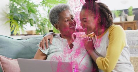 Grandmother and granddaughter sharing laptop, smiling and embracing on sunlit sofa