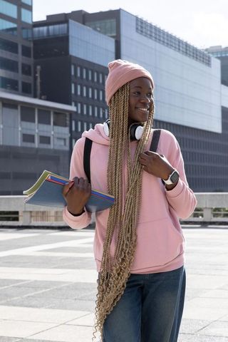 African american student in pink hoodie and beanie with long blonde braids carrying notebooks