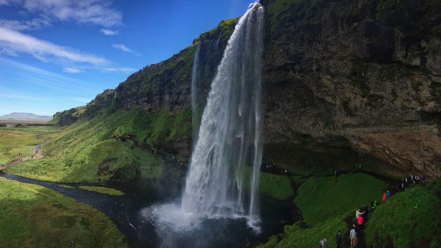 Tourists are standing near and admiring a tall, cascading waterfall in Iceland, surrounded by lush green vegetation and a rocky cliff. The sky is clear, and the water creates a mist around the base of the waterfall. Perfect for use in travel brochures, nature-themed articles, adventure promotion, scenic landscape calendars, and environmental conservation campaigns.