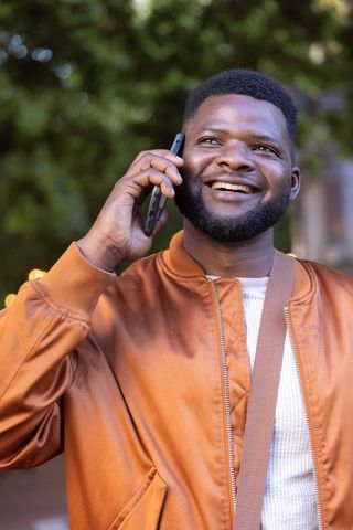 Smiling african man talking on phone outdoors wearing bomber jacket and crossbody bag