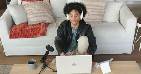 African american man sitting on floor wearing headphones using laptop and mic for podcasting