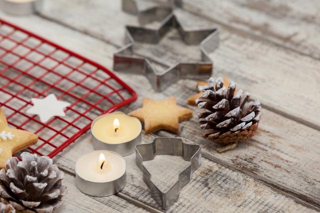 This image showcases a rustic holiday scene with Christmas-themed cookie cutters, pine cones, and tealight candles on a wooden table. Ideal for use in holiday baking promotions, festive decoration ideas, or seasonal greeting cards.