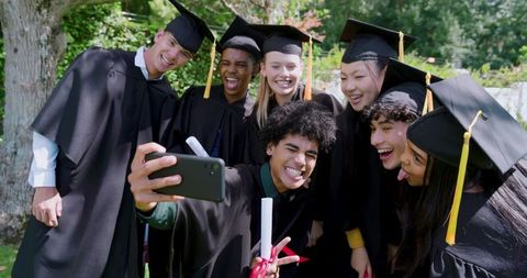 Diverse Graduates Taking Selfie on Campus Lawn Wearing Caps and Gowns Holding Diplomas