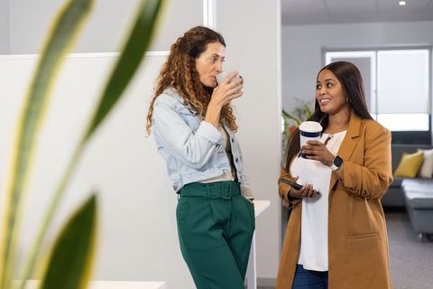 Two professional women chatting and drinking coffee during break in modern office lounge
