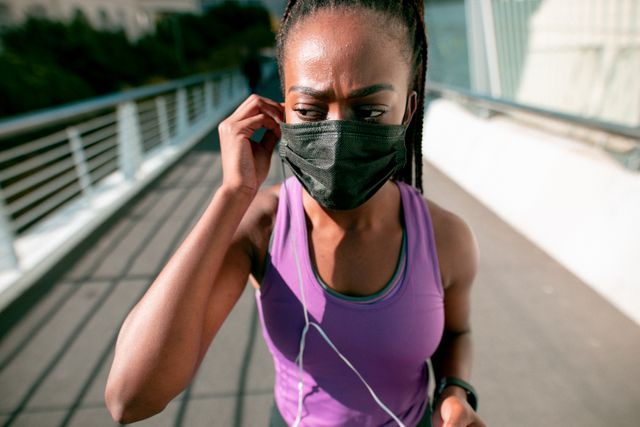 African American woman wearing a face mask while jogging in an urban environment. Ideal for themes related to health, fitness, COVID-19 safety measures, and outdoor activities. Can be used in articles promoting healthy living during the pandemic, fitness blogs, and public health campaigns.