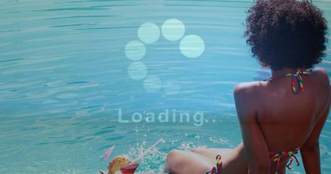 Woman in colorful string bikini sitting by turquoise water, splashing toes with tropical cocktail