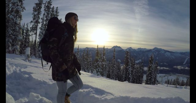 Young woman hiking in snowy mountain landscape at sunset, wearing winter gear and backpack. Snow-covered trees and mountains create a serene scene. Useful for themes related to adventure travel, winter sports, outdoor activities, scenic nature, and exploration.