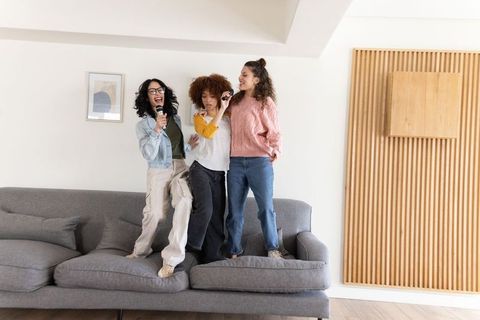 Three diverse friends standing on sofa singing karaoke together in modern living room