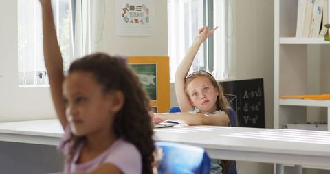Young student raising hand while listening in bright classroom with sunlight and blinds