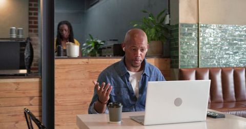 Man gesturing during video call at coffee shop with laptop and takeaway coffee