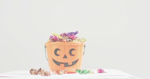 Jack-o'-lantern candy pail overflowing with colorful foil-wrapped sweets on white tabletop