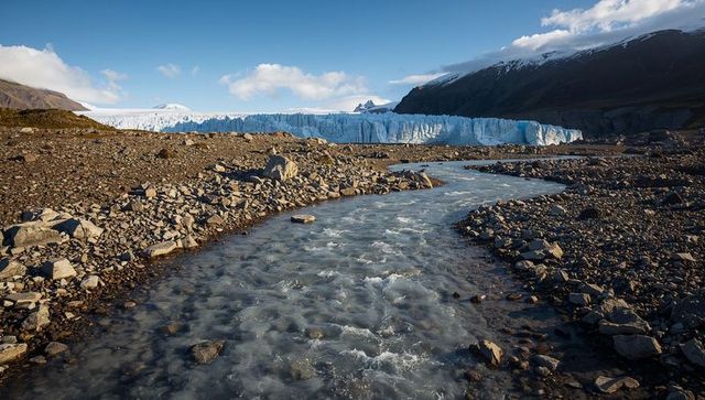 Flowing glacial meltwater river carving rocky foreland toward blue-ice glacier panorama