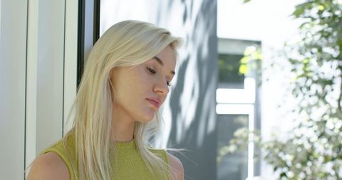 Blonde woman standing by glass door wearing green knit tank top in sunlit modern interior