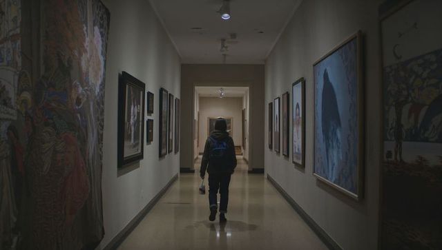 Solitary figure walking down dim museum corridor lined with framed paintings and tapestry