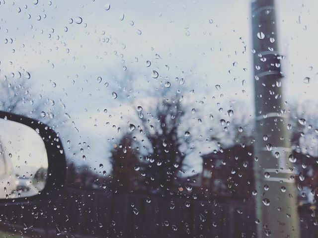 Raindrops collecting on a car window obscuring an outdoor scene of trees and houses. Use for themes around weather, rain, travel, nature, or melancholy settings.