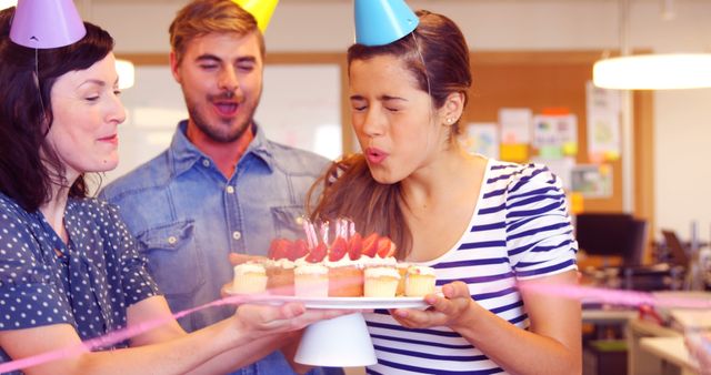 Woman blowing out candles on cake surrounded by smiling coworkers during office birthday celebration. The scene includes party hats, balloons, and a cheerful environment. Perfect for promoting workplace unity, celebrating employee milestones, or emphasizing a fun and supportive work environment.