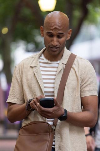 Urban man checking smartphone on tree-lined sidewalk with leather crossbody bag